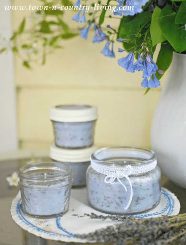 Three glass jars filled with lavender lotion rest on a doily-covered table, accompanied by sprigs of lavender and vibrant blue flowers in a white vase, creating an ambiance reminiscent of soothing lavender scented candles.